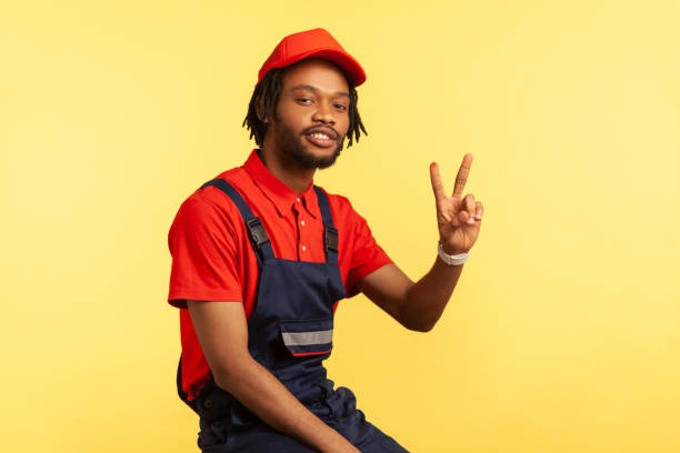 istock Handyman wearing blue uniform, red visor cap and T-shirt posing, looking at camera, showing v sign. 1348686486
