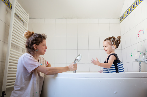toddler boy, almost three years old, wearing bathing suit with built-in swim belt with blue and white stripes, has been finger painting in bathroom, some color is still on the wall, now, with the help of his mother, he is taking a shower to wash of the color
