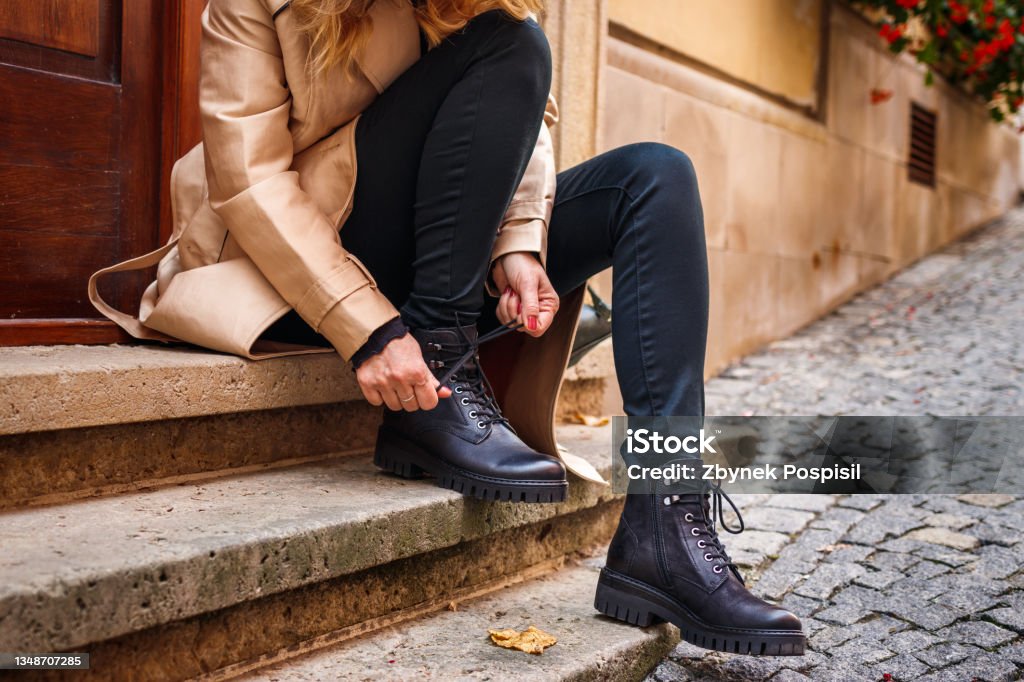 Stylish woman tying shoelace of black ankle boot Woman wearing trench coat sitting on staircase and tying shoelace on her ankle boot. Autumn fashion collection. Trendy black leather shoe. Street style Boot Stock Photo