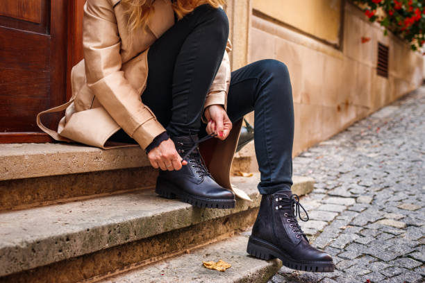 Stylish woman tying shoelace of black ankle boot Woman wearing trench coat sitting on staircase and tying shoelace on her ankle boot. Autumn fashion collection. Trendy black leather shoe. Street style boot stock pictures, royalty-free photos & images