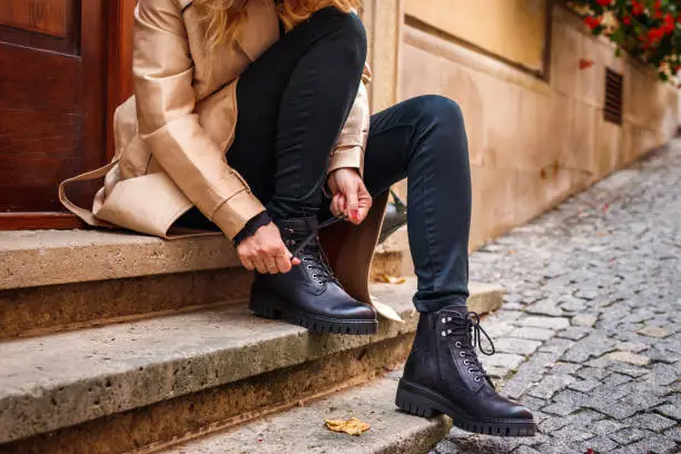 Woman wearing trench coat sitting on staircase and tying shoelace on her ankle boot. Autumn fashion collection. Trendy black leather shoe. Street style