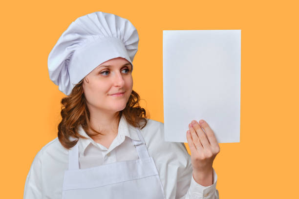 istock Happy woman chef with blank A4 paper sheet for text in hand on studio background, mockup copy space 1348717033