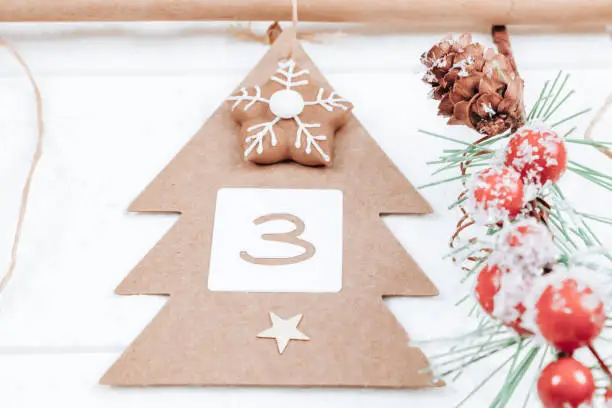 One handicraft Christmas tree with number 3, biscuits and a spruce branch hang on a jute thread and on a hanger on a white brick wall, bottom close-up view. Advent calendar concept.