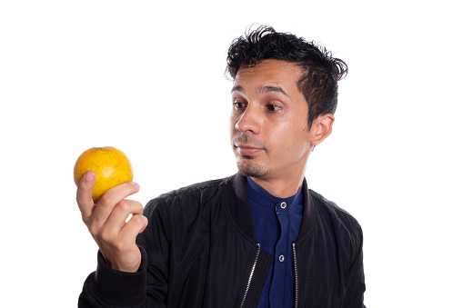 Man holding and observing an orange fruit. White background. Young latin man looking at appetizing and delicious orange fruit.