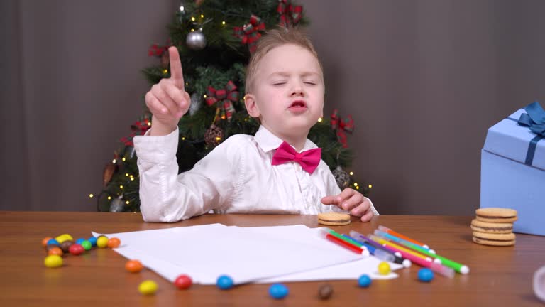 Spoiled boy does not want to draw postcard, write letter with wishes to Santa or does homework so he indulges, talks and eats candy, decorated Christmas tree on background