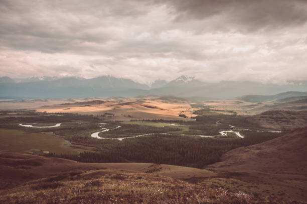 paysage vintage avec vaste plateau avec rivière de montagne et forêt sur fond de crête de montagne enneigée sous un ciel nuageux. vallée de montagne et chaîne de montagnes parmi les nuages bas dans des tons sépia et délavés. - russia river landscape mountain range photos et images de collection