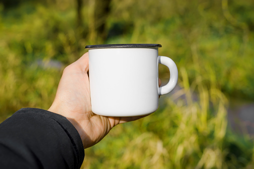 Metal mug mock-up, outdoors. Close-up of man's hand holding empty, hiking enamelled cup in forest against background of green grass.