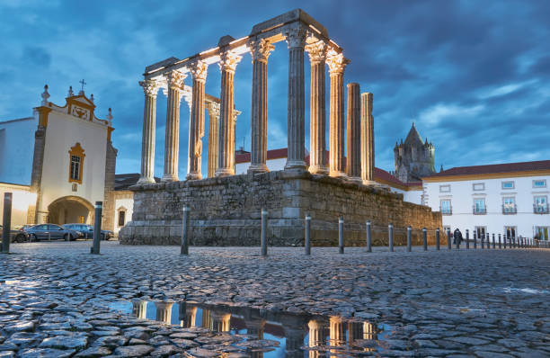 Roman temple ruins with illuminated columns at twilight, reflected in rain-wet cobblestone plaza.