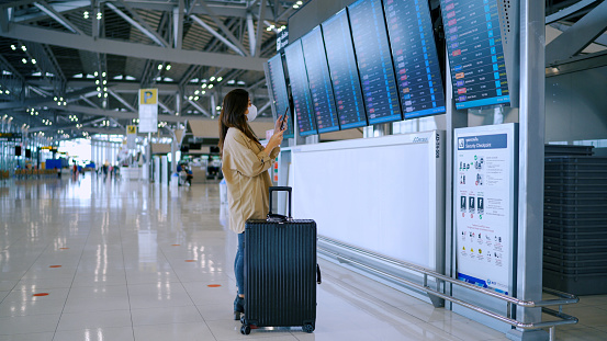 Asian business woman wear surgical mask checking boarding time at digital timetable at the airport terminal.