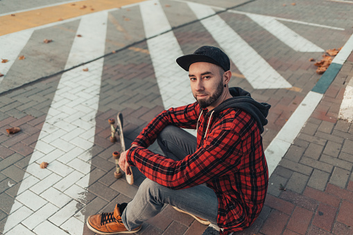 Skateboarding. A young man in a black cap, jeans and a red plaid shirt sits on a skateboard. The view from the top.