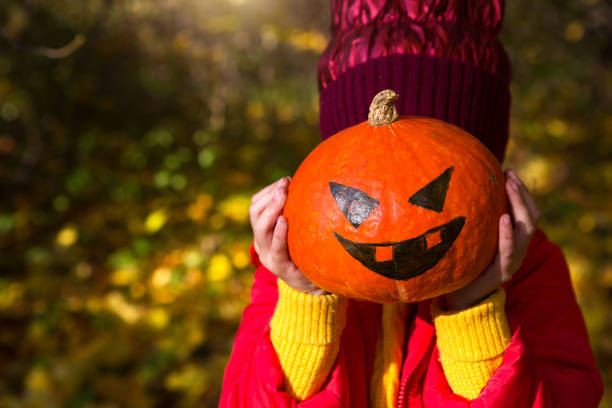 une fille vêtue de vêtements chauds rouges et jaunes tient une citrouille avec des yeux et une bouche peints dans les mains. halloween, jack lantern, sans visage. humeur automnale, feuilles sèches jaunes - child autumn scarecrow decoration photos et images de collection