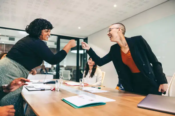 Cheery businesswomen fist bumping each other before a meeting in a boardroom. Two colleagues smiling cheerfully while greeting each other. Group of businesspeople attending a briefing in a modern office.