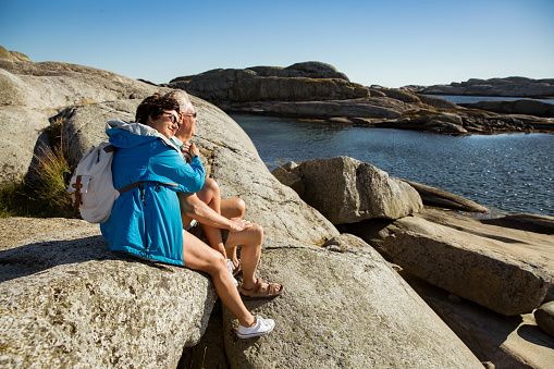 Loving mature couple traveling, sitting on the rock, exploring. Real man and woman hugging, kissing, Happily smiling. Scenic view of Serene Scandinavian landscape on south coast of Norway, Tjøme.