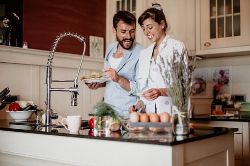 Beautiful loving couple making breakfast together. A man in a denim shirt and a woman in a white shirt are standing above the sink in the kitchen and making eggs and vegetables together.