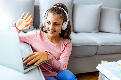 Happy little female teenager in headphones looking at laptop screen, waving hand making hello gesture, starting online video web camera zoom call with teacher or friends, distant communication concept