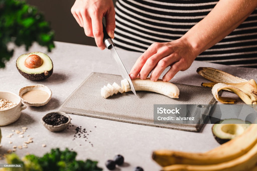 Woman chopping banana for making smoothie bowl in kitchen Close-up of a woman cutting banana on chopping board with black sesame seeds, tahini, avocado on kitchen counter.  Female preparing smoothie bowl at home kitchen. Banana Stock Photo