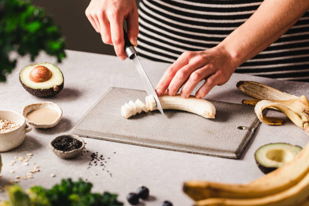 Woman chopping banana for making smoothie bowl in kitchen Close-up of a woman cutting banana on chopping board with black sesame seeds, tahini, avocado on kitchen counter. Female preparing smoothie bowl at home kitchen. chopping food stock pictures, royalty-free photos & images