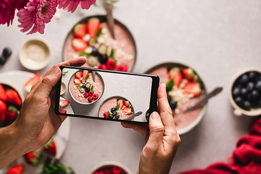 Close-up of a woman photographing strawberry smoothie bowls on the kitchen counter. Hands of a female taking photo of fruit smoothie bowl with her mobile phone.
