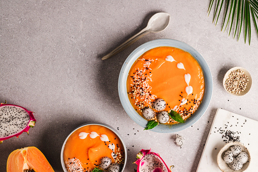 Directly above shot of papaya and mango smoothie bowl garnished with cream, dragon fruit balls, and mint leaves on the kitchen counter. Healthy fruit smoothie bowl with ingredients on kitchen counter.