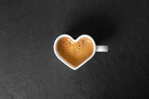 white cup in the shape of a heart with coffee and beautiful foam on a black stone background. view from above. copy space. High quality photo