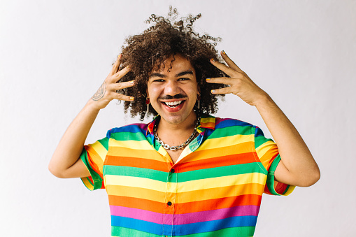 Smiling young gay man embracing his identity in a studio. Young man looking at the camera cheerfully while standing against a white background. Young gay man wearing a shirt with rainbow colours.