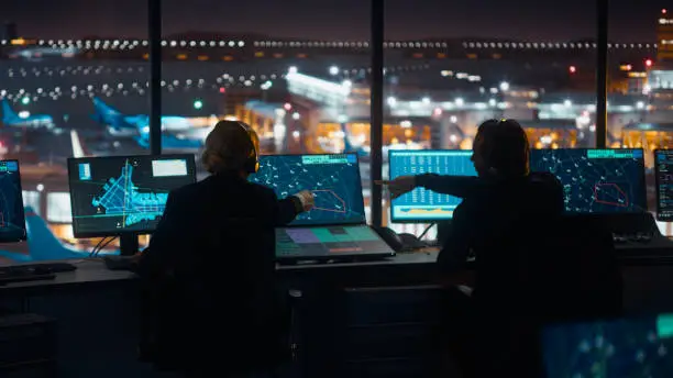 Photo of Diverse Air Traffic Control Team Working in a Modern Airport Tower at Night. Office Room is Full of Desktop Computer Displays with Navigation Screens, Airplane Flight Radar Data for Controllers.