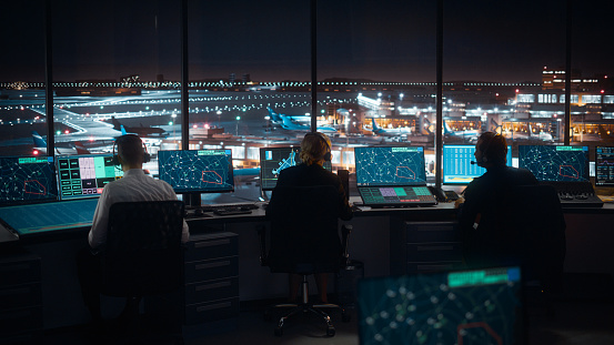 Diverso equipo de control de tráfico aéreo que trabaja en una moderna torre de aeropuerto por la noche. La sala de oficina está llena de pantallas de computadoras de escritorio con pantallas de navegación, datos de radar de vuelo de avión para control photo