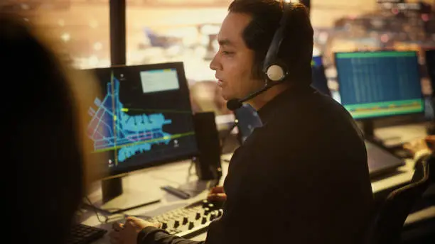 Photo of Portrait of Male Air Traffic Controller with Headset Talk on a Call in Airport Tower. Office Room is Full of Desktop Computer Displays with Navigation Screens, Airplane Flight Radar Data.