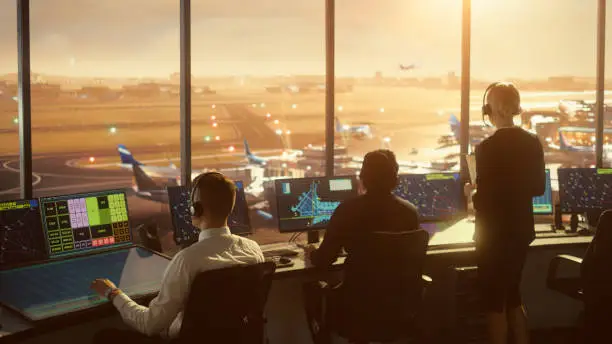 Photo of Diverse Air Traffic Control Team Working in a Modern Airport Tower at Sunset. Office Room is Full of Desktop Computer Displays with Navigation Screens, Airplane Flight Radar Data for Controllers.