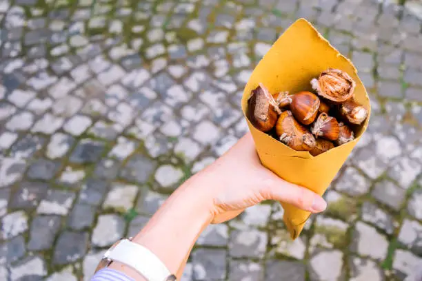 A woman's hand with a white clock holds a yellow paper cornet of roasted chestnuts against the gray background of street cobblestones