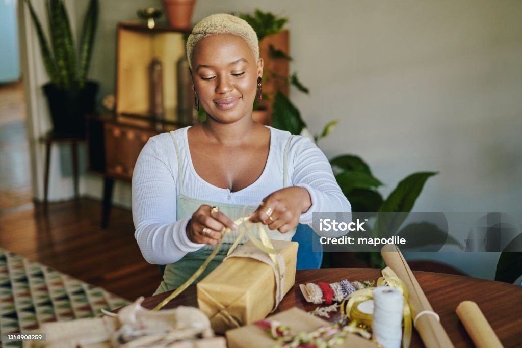 Smiling young woman wrapping Christmas presents with recycled paper Smiling young woman sitting at a table at home and using recycled paper and ribbons to wrap Christmas presents for the holidays Gift Stock Photo