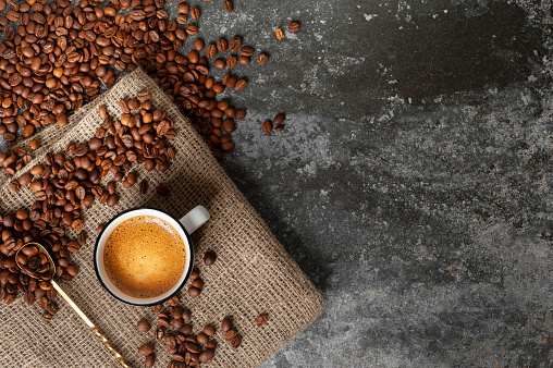 a cup of coffee with a beautiful froth stands on a fabric against a concrete background, coffee beans are scattered nearby. copy space