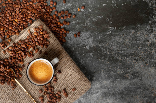a cup of coffee with a beautiful froth stands on a fabric against a concrete background, coffee beans are scattered nearby. copy space - coffee top view imagens e fotografias de stock