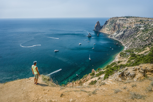 Man traveler enjoying landscape of Cape Fiolent locate in Crimea. Sea view - Mountains surround the bay and the small pleasure yacht boats moored in the lagoon.