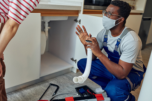 Black handyman in overalls installing a kitchen sink on an customer’s home. He is wearing protective face mask