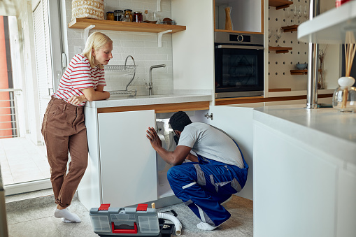 Black male plumber in uniform fixing kitchen sink. He is squatting and solving the problem
