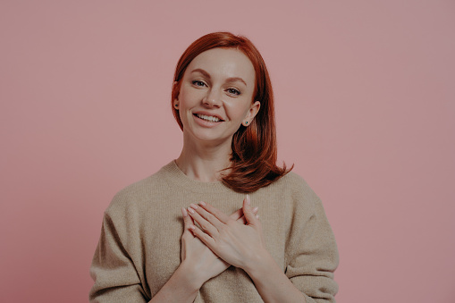 Wishful peaceful young red-haired woman holding hands on chest and thanking, showing gratitude sign while standing against pink wall in studio, thankful ginger female looking gratefully at camera