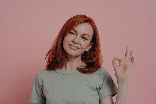 Body language concept. Calm satisfied redhead female showing OK sign, making okay gesture with fingers while posing isolated over pink studio background, smiling woman giving her approval