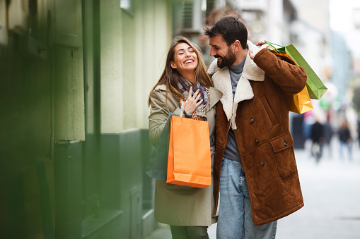 A young woman is smiling and enjoying shopping with her boyfriend. They are holding shopping bags and walking down the street, enjoying Autumn day.