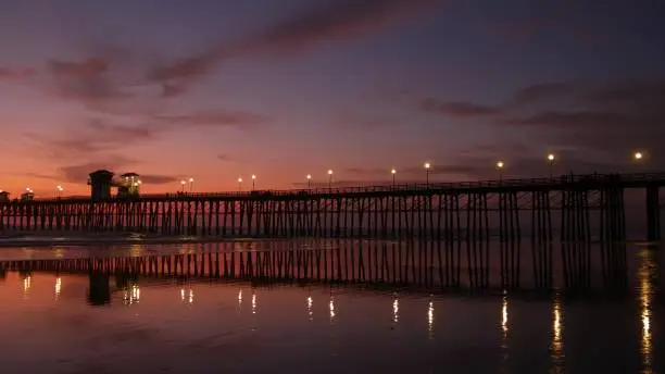 Pier silhouette Oceanside California USA. Pacific ocean tide tropical beach. Summertime gloaming atmosphere. Purple aesthetic gradient, calm twilight sky, pink violet dusk. Lights reflection in water.