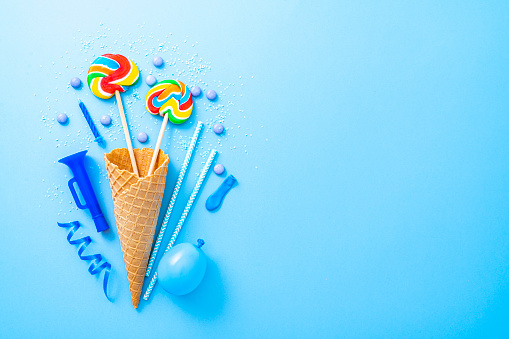 Party, birthday or carnival backgrounds. Overhead view of blue colored accessories like balloons, drinking straws, confetti, candies, lollipop, ice cream cone and streamers shot at the left of a blue background leaving useful copy space for text and/or logo. High resolution 42Mp studio digital capture taken with SONY A7rII and Zeiss Batis 40mm F2.0 CF lens