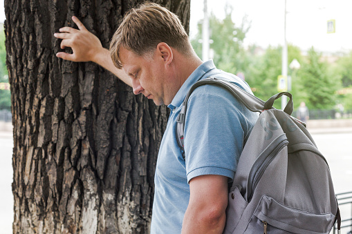 Un hombre adulto con una mochila apoyó su mano en un árbol sintiéndose enfermo en la calle photo