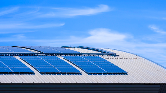 Many solar panels on top of curved steel roof of industrial building against blue sky background