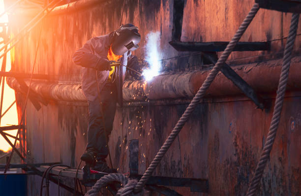 welder in protective workwear on scaffold is welding the old rusty vessel hull in shipyard area at sunset time