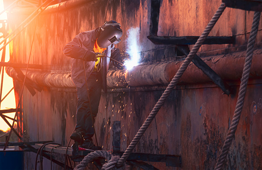 Side view of welder in protective workwear on platform is welding the old rusty vessel hull in shipyard area at sunset time