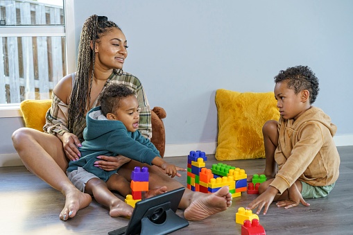 A beautiful young black mother sits on the floor at home with her two young sons. She is holding one of the boys in her lap. The children are having a disagreement and the woman is patiently mediating. The kids have been playing with colorful plastic toy building blocks. A tablet computer rests on the floor in front of the family.