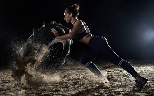 спортсменка переворачивает колесо шин в спортзале, распыляя песок. концептуальная тренировка, рвение к победе, преодоление себя. женщина т� - health club фотографии стоковые фото и изображения