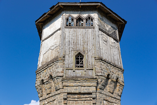 The upper part of an ancient watchtower made of stone blocks and wood paneling with windows against the blue sky on a bright sunny summer day. Close-up