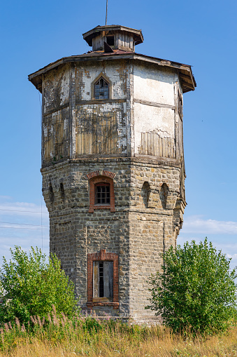 Old stone water tower with windows and a wooden roof against the background of blue sky and green trees on a sunny summer day. Close-up