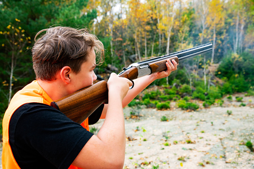 A young male hunter firing a shotgun outside in the woods on an autumn day.